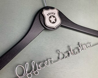 Puede incluir: Una percha de madera negra con una placa de policía plateada que dice "POLICE" con una estrella en el centro. La percha tiene una inscripción de alambre plateado que dice "Officer Salvatore".