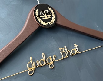 May include: A brown wooden hanger with a black circle featuring a gold scale of justice and laurel wreath. The hanger has a gold wire inscription that reads "Judge Bhat".