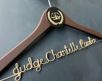 May include: A brown wooden hanger with a gold wire inscription that reads "Judge Chartelle Carter". The hanger has a black circle with a gold scale of justice and laurel wreath design.