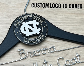 May include: A black wooden clothes hanger with a silver metal hook. A circular black plaque features the white UNC Medical School logo and the text October 2024. Below, silver wire is bent to spell out the name Brenna and 1st White Coat.