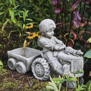 May include: A grey concrete statue of a child driving a toy tractor with a wagon attached. The child is wearing a baseball cap and has a dog sitting on the tractor with them.