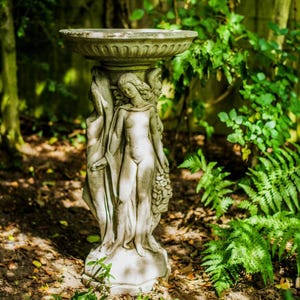 May include: A stone bird bath with a sculpted design of three figures. The bird bath is in a garden setting with green foliage.