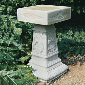 May include: A light grey stone bird bath with a square basin and a decorative pedestal. The basin has diamond-shaped carvings. The pedestal features relief carvings of figures and floral designs. The bird bath is surrounded by green foliage.