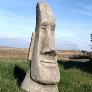 May include: A large stone sculpture of a human face with a closed-mouth smile. The sculpture is weathered and has a rough texture. The sculpture is standing on a grassy field.
