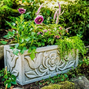 May include: A rectangular concrete planter with an ornate floral design. The planter is filled with green plants and pink flowers.