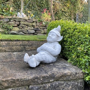 May include: A grey concrete garden gnome statue sitting on a stone step. The gnome is wearing a pointed hat and has a relaxed expression on its face. The gnome is sitting with its legs crossed and its hands resting on its lap.