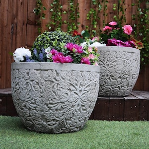 May include: Two grey concrete planters with intricate floral designs, filled with pink and white flowers, and green foliage. The planters are sitting on a wooden deck.