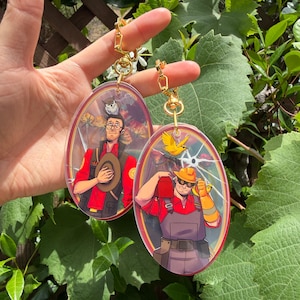 Op de afbeelding: Twee ovale acryl sleutelhangers met Team Fortress 2-personages. De ene toont een figuur in een rood shirt met een hoed en een uil, de andere een figuur met een helm en overall die een rode gereedschapskist en een gele vogel bij zich draagt.