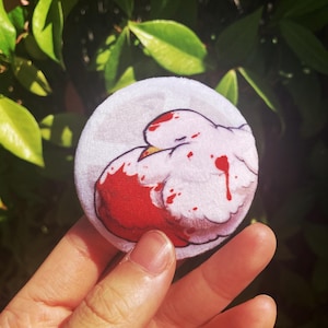 Pode incluir: Um patch redondo de pelúcia branca com um desenho de pássaro de desenho animado. O pássaro tem uma asa vermelha e gotas vermelhas semelhantes a sangue. O patch é segurado em uma mão contra um fundo de folhas verdes.