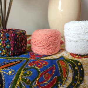 May include: Three yarn skeins in a row. A multi-colored, textured yarn cube sits to the left. A peach-colored yarn ball is in the middle. A white, fluffy yarn ball is on the right. The yarns are on a blue and cream patterned surface.
