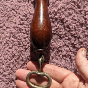 May include: A wooden handle with a metal ring attached to the bottom. The handle is dark brown and has a smooth finish. The ring is silver and has a simple design.