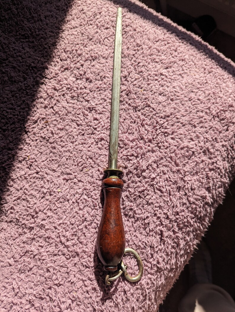 May include: A metal knife sharpener with a wooden handle and a metal ring attached. The sharpener is on a pink fuzzy surface.