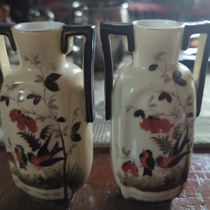 May include: Two small, white ceramic vases with black handles and floral designs. The vases feature a bird and floral pattern in shades of red, black, and gray.