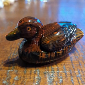 Può includere: Una statuetta di anatra di legno con finitura dipinta marrone, nero e arancio. L'anatra è seduta su una superficie di legno.