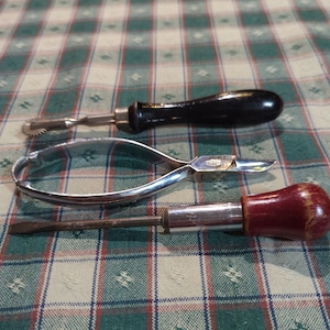 May include: Three tools on a checkered tablecloth: a small, silver tool with a wheel, a pair of silver pliers, and a screwdriver with a red handle.