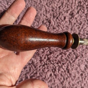 May include: A close-up of a wooden handled sharpening steel with a metal blade. The handle is dark brown and has a smooth finish. The metal blade is silver and has a rough texture.