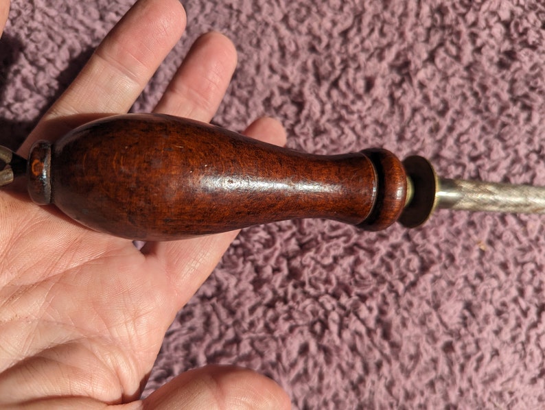 May include: A close-up of a wooden handled sharpening steel with a metal blade. The handle is dark brown and has a smooth finish. The metal blade is silver and has a rough texture.