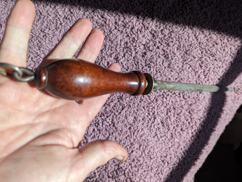 May include: A brown wooden handled steel knife sharpener with a metal ring and a chain attached to the handle.
