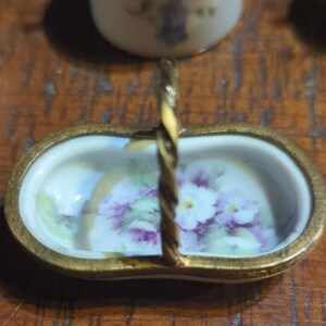 May include: A small, white porcelain basket with a gold rim and a twisted gold handle. The basket is decorated with a floral pattern of purple and white flowers.
