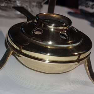 May include: A gold metal tea warmer with a round top and three legs. The warmer has a small hole in the centre and several smaller holes around the edge.