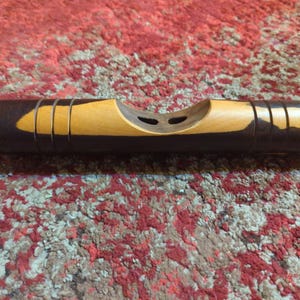 Puede incluir: Un nivel de madera hecho a mano con una combinación de colores marrón oscuro y marrón claro. El nivel tiene una parte superior curvada con dos agujeros y varias ranuras verticales. El nivel mide unos 20 cm de largo.