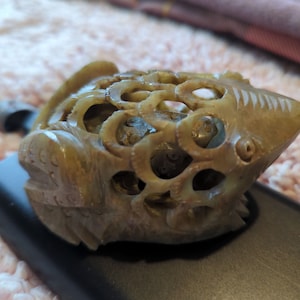 Op de afbeelding: Een groene jade gegraveerde kikkerfiguur met ingewikkelde details en een uitgeholde binnenkant. De kikker zit op een zwart oppervlak.