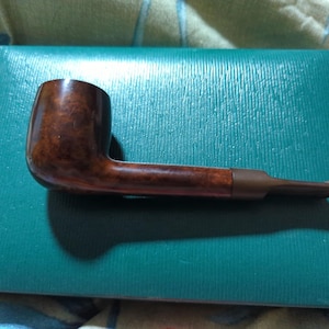 May include: A wooden smoking pipe in shades of brown, featuring a curved bowl and a long stem. The pipe is displayed on a teal-coloured, textured surface.