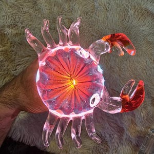 May include: A clear glass crab sculpture with red and orange detailing. The crab's body features a red, flower-like design in the centre, and the claws are bright red. The sculpture is illuminated from within, creating a glowing effect.