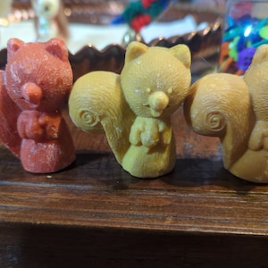 May include: Three small squirrel figurines, one red, one yellow, and one brown, with a wood grain background.