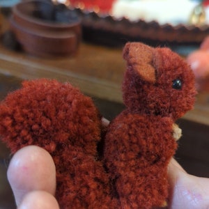 Può includere: Un piccolo giocattolo di scoiattolo rosso e peloso con una lunga coda folta. Il giocattolo è fatto di filato e ha un viso carino e da cartone animato.