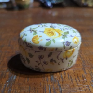 Könnte beinhalten: Eine kleine, runde, weiße Keramik-Schmuckdose mit Blumenmuster. Die Dose ist mit gelben Rosen und lila Blumen auf weißem Hintergrund verziert.