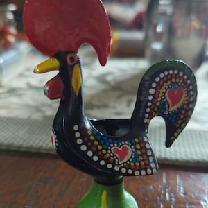 以下が含まれることがあります： 赤い鶏冠と緑色の台座が付いた黒い雄鶏の置物。雄鶏は白、黄、赤、青のドットとハートで飾られています。