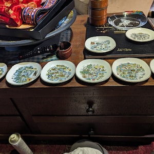 May include: Seven small, white ceramic plates with gold trim. Each plate features a colourful illustration of a city map with buildings and landmarks. The plates are arranged in a row on a wooden surface.