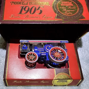 Puede incluir: Un modelo en miniatura de locomotora de vapor azul y roja, presentado en una caja roja. El modelo presenta ruedas doradas y está etiquetado como "1905 Fowler Showmans Engine". La caja está marcada como "Models of Yesteryear".