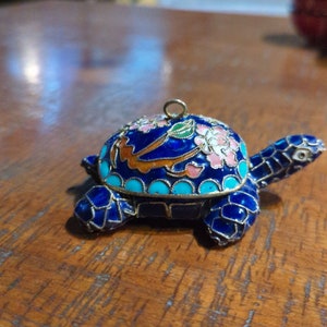 Puede incluir: Un adorno de tortuga de cloisonné azul y dorado con detalles florales. La tortuga está hecha de metal y tiene un acabado brillante.