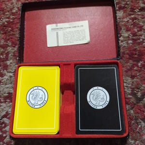 Könnte beinhalten: Vintage Spielkarten-Set in einem rot-schwarzen Etui. Die Karten sind gelb und schwarz mit einem weißen Logo mit der Aufschrift "Milton Pipes". Das Etui enthält einen Papiereinleger mit Text von "Waddington's Playing Card Co Ltd".