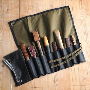 Peut inclure: Un rouleau à outils ouvert avec divers outils de menuiserie. Le rouleau est vert olive avec une bordure noire et une poche zippée noire. Les outils comprennent des ciseaux et d'autres outils à manche en bois. Les outils sont exposés sur une surface en bois.