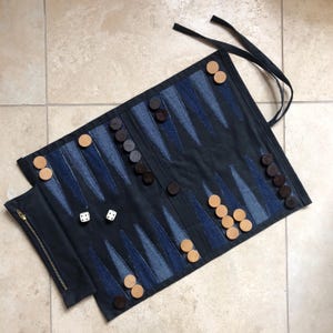 May include: A rolled-up backgammon board with a dark blue denim design and a zippered pouch. The board features blue and black triangles, wooden game pieces, and two dice. The game is ready to play.