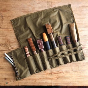 May include: An olive green tool roll containing various woodworking tools. The tools include chisels and other hand tools with wooden handles and leather or wrapped grips. The roll is open, displaying the tools in individual pockets, and has a zipped pocket on the side.