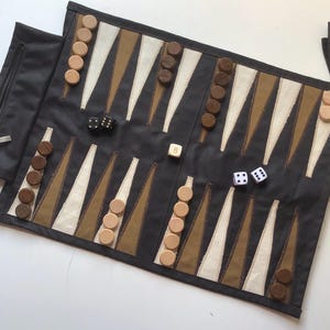 May include: A rolled-up backgammon game set. The game board is black with tan and white triangles. Wooden game pieces are light and dark brown. Two black dice, one white die with the number 8, and two white dice with the numbers 1 and 2 are visible. The set includes a zippered pouch.