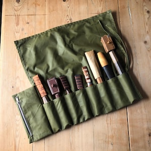 Puede incluir: Un rollo de herramientas de lona verde oliva con cremallera, que contiene una variedad de herramientas de tallado de madera. Las herramientas tienen diferentes formas y colores de mango, incluyendo marrón, negro y tonos de madera natural. El rollo está abierto sobre una superficie de madera.