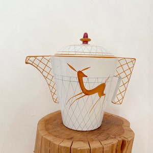 Könnte beinhalten: Weiße Teekanne aus Keramik mit geometrischem Muster und einem stilisierten orangefarbenen Hirsch-Design. Die Teekanne hat einen Deckel mit einem kleinen roten Knopf und zwei Griffen.