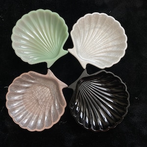 May include: Four seashell-shaped dishes in pastel colors: light green, white, pink, and black. The dishes have a ribbed texture and are arranged on a dark background. These decorative dishes are ideal for serving small portions.