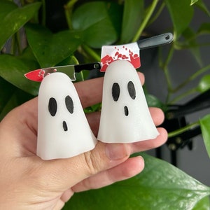 May include: Two white ghost-shaped decorations with black eyes and mouths. Above them is a small knife with a red blade and blood splatter design. The decorations are held in front of a background of green leaves.