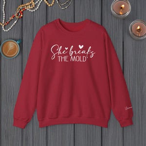 May include: Red sweatshirt with the text "She breaks the mold" in white with two small hearts above the word "breaks".
