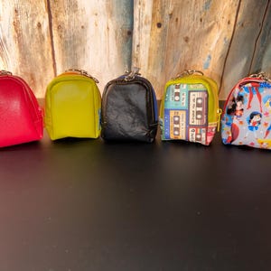 May include: Five miniature backpack-shaped coin purses in various colors and patterns. The purses include red, yellow, black, a cassette tape design, and a Wonder Woman print. Each has a zipper closure and a small metal loop.