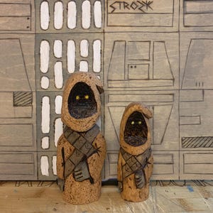 Può includere: Due figure in legno intagliate di personaggi di Star Wars, un Jawa e un Tusken Raider, in piedi davanti a uno sfondo in legno con un motivo di quadrati e rettangoli.