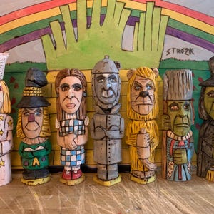 Può includere: Sette figurine di legno che rappresentano i personaggi de Il Mago di Oz. I personaggi sono Dorothy, lo Spaventapasseri, l'Uomo di Latta, il Leone Codardo, la Strega Cattiva dell'Ovest e il Mago. Le figurine sono dipinte con colori vivaci e hanno dettagli elaborati. Lo sfondo è un fondale dipinto di verde e giallo con un arcobaleno.