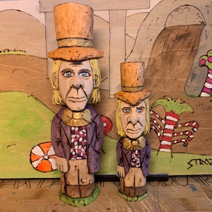 Può includere: Due figure di legno intagliate di un uomo con cappello a cilindro, cappotto viola e camicia fantasia. Le figure sono davanti a uno sfondo di legno dipinto con un paesaggio stravagante. La figura più grande è alta circa 25 cm.