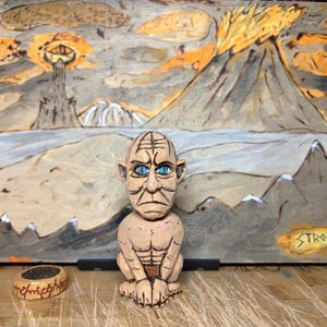 Può includere: Una scultura in legno di una creatura con occhi blu e un'espressione severa, posta davanti a un dipinto di paesaggio. Un anello con testo è in primo piano. Il dipinto raffigura una catena montuosa e una torre con un occhio.